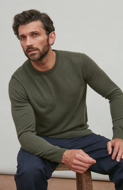 Man wearing a green merino cashmere crew neck sweater and dark pants sitting on a wooden stool against a plain background