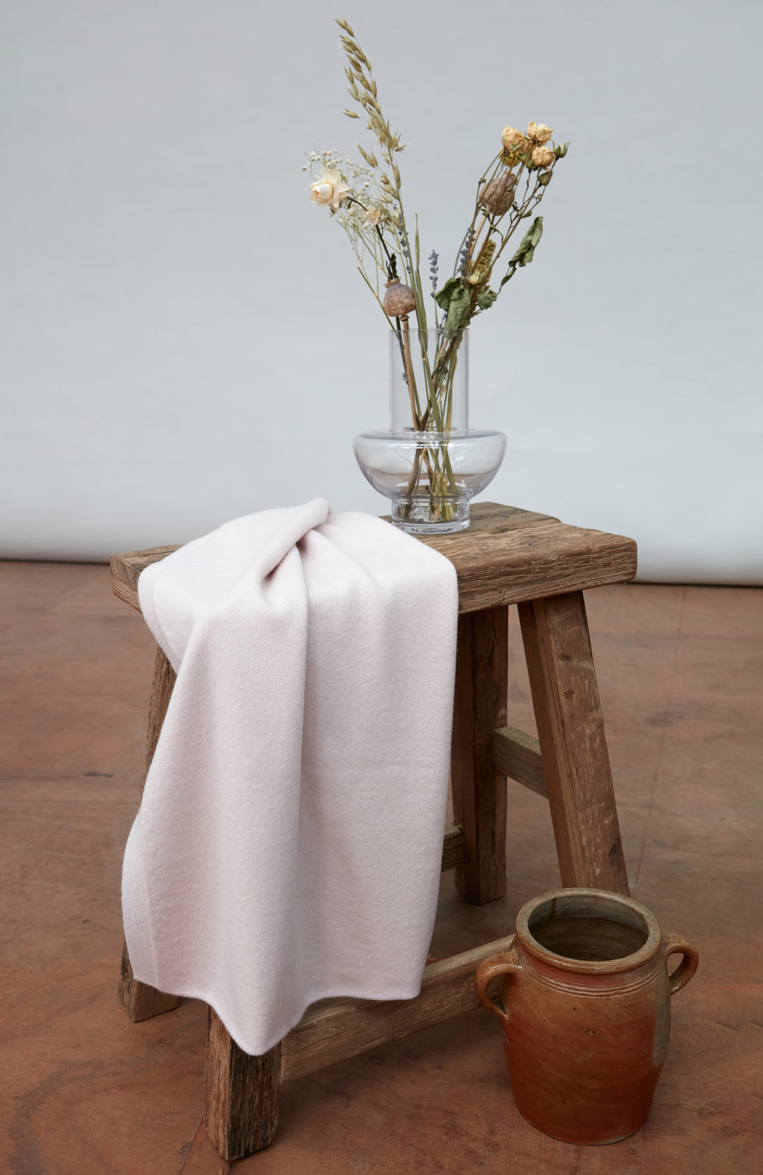 A pale blush pink knit light cream merino cashmere shawl draped over a rustic wooden stool, with a glass vase of dried flowers and a brown ceramic pot on the floor against a neutral studio backdrop.