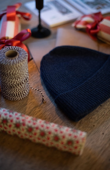 navy merino cashmere knit beanie, spool of twine, and roll of patterned paper on a wooden surface.