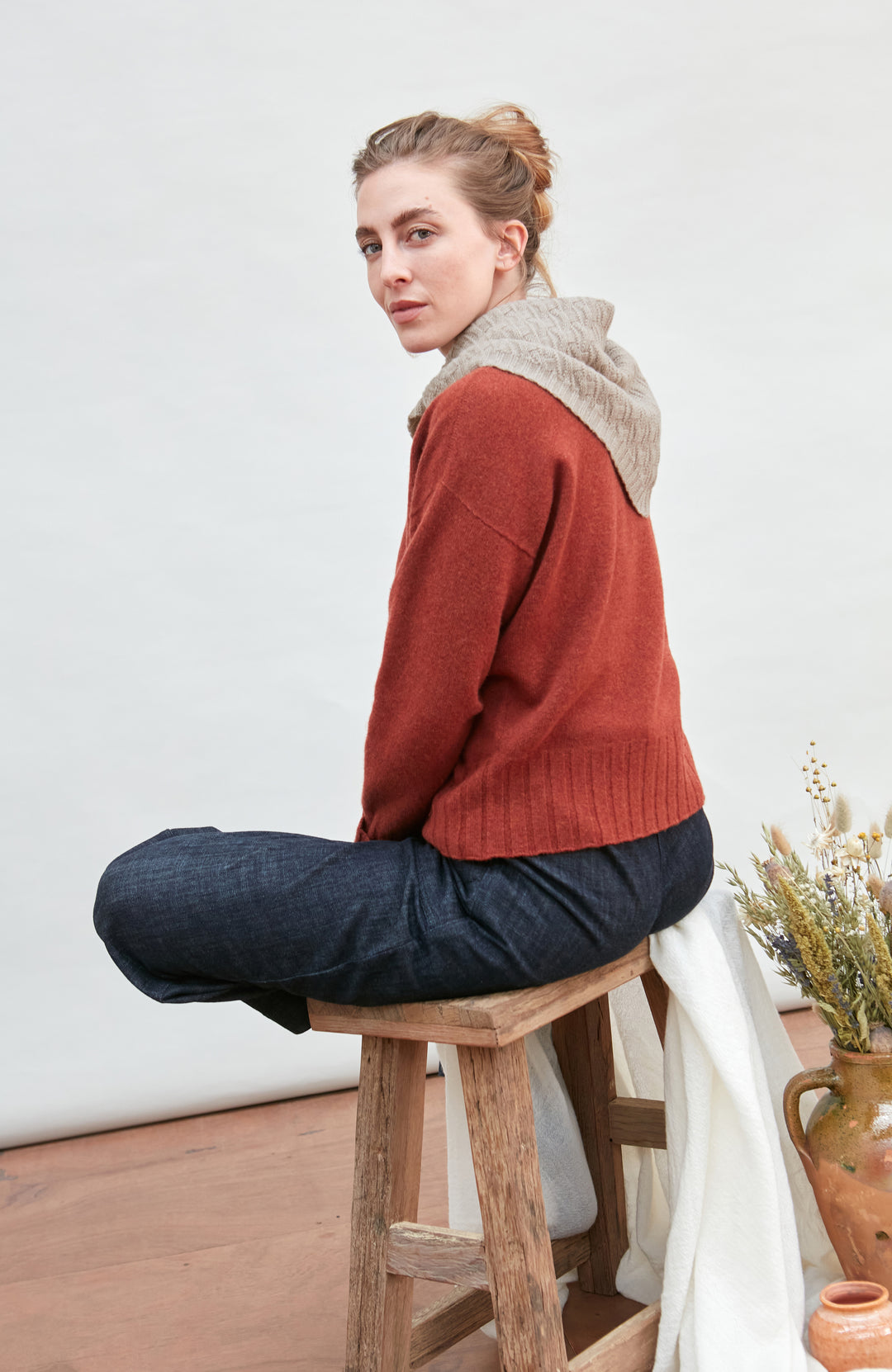 Woman sitting on a wooden stool wearing a beige merino cashere knitted neckerchief with a white wall and decorative elements in the background by Paul James