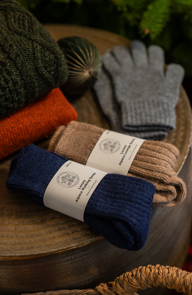 Pair of blue alpaca socks with a label, brown socks, and grey gloves on a woven surface with Christmas decor in the background.