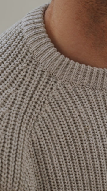 Man sitting on a chair wearing a light gray cotton fisherman sweater by Paul James Knitwear and dark jeans against a plain background